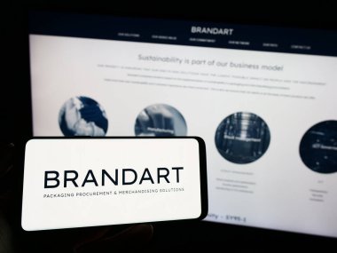 Stuttgart, Germany - 09-20-2023: Person holding mobile phone with logo of Italian packaging company Brandart  S.p.A. on screen in front of web page. Focus on phone display. clipart