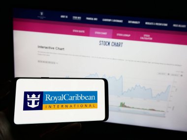 Stuttgart, Germany - 08-07-2023: Person holding mobile phone with logo of company Royal Caribbean International (RCI) on screen in front of web page. Focus on phone display. clipart