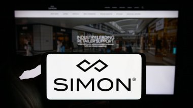 Stuttgart, Germany - 08-08-2023: Person holding smartphone with logo of US real estate company Simon Property Group Inc. on screen in front of website. Focus on phone display. clipart