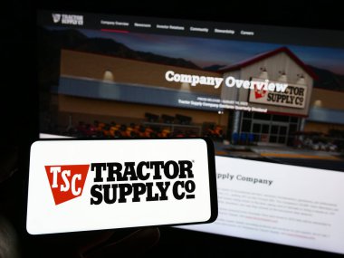 Stuttgart, Germany - 08-11-2023: Person holding smartphone with logo of US retail business Tractor Supply Company on screen in front of website. Focus on phone display. clipart