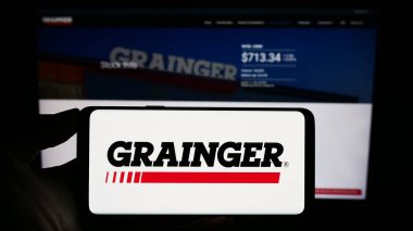 Stuttgart, Germany - 08-12-2023: Person holding cellphone with logo of US industrial supply company W.W. Grainger Inc. on screen in front of webpage. Focus on phone display. clipart