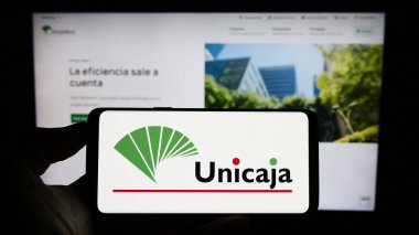 Stuttgart, Almanya - 08-13-2023: İspanyol bankacılık şirketi Unicaja Banco SA 'nın logosuyla cep telefonu sahibi kişi iş sayfasının önünde ekranda. Telefon ekranına odaklan.