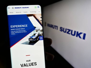 Stuttgart, Germany - 08-14-2023: Person holding cellphone with webpage of Indian company Maruti Suzuki India Limited on screen in front of logo. Focus on center of phone display. clipart