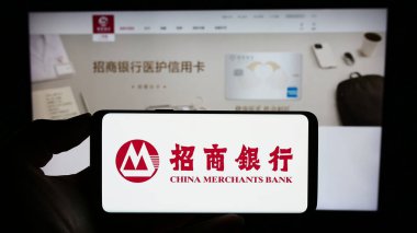 Stuttgart, Germany - 08-14-2023: Person holding mobile phone with logo of China Merchants Bank Company Limited (CMB) on screen in front of web page. Focus on phone display. clipart