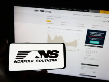 Stuttgart, Germany - 08-14-2023: Person holding mobile phone with logo of American railway company Norfolk Southern Corp. on screen in front of web page. Focus on phone display. clipart