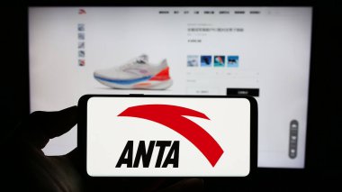 Stuttgart, Germany - 08-15-2023: Person holding cellphone with logo of Chinese company Anta Sports Products Limited on screen in front of webpage. Focus on phone display. clipart