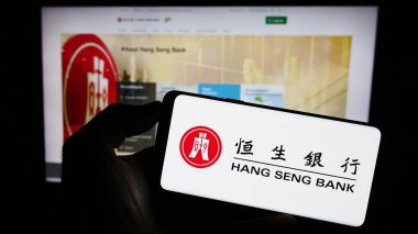 Stuttgart, Germany - 08-16-2023: Person holding mobile phone with logo of Chinese company Hang Seng Bank Limited on screen in front of business web page. Focus on phone display. clipart