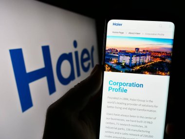 Stuttgart, Germany - 08-17-2023: Person holding cellphone with webpage of home appliances company Haier Group Corporation on screen with logo. Focus on center of phone display. clipart