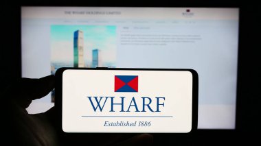 Stuttgart, Almanya - 08-17-2023: Çinli şirket The Wharf Holdings Limited 'in logosuyla cep telefonu sahibi kişi iş sayfasının önünde ekranda. Telefon ekranına odaklan.