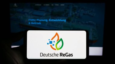 Stuttgart, Germany - 08-20-2023: Person holding mobile phone with logo of German company Deutsche ReGas GmbH Co. KGaA on screen in front of web page. Focus on phone display. clipart