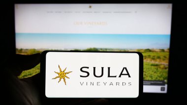 Stuttgart, Germany - 08-20-2023: Person holding mobile phone with logo of Indian winery company Sula Vineyards on screen in front of business web page. Focus on phone display. clipart