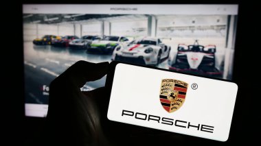 Stuttgart, Germany - 08-20-2023: Person holding smartphone with logo of automotive company Dr. Ing. h.c. F. Porsche AG on screen in front of website. Focus on phone display. clipart