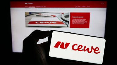 Stuttgart, Germany - 08-21-2023: Person holding mobile phone with logo of printing company CEWE Stiftung Co. KGaA on screen in front of business web page. Focus on phone display. clipart