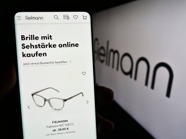 Stuttgart, Almanya - 08-21-2023: Alman gözlük şirketi Fielmann AG 'nin web sayfası ile logo önünde akıllı telefon tutan kişi. Telefon ekranının merkezine odaklan.