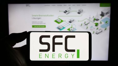 Stuttgart, Germany - 08-22-2023: Person holding smartphone with logo of German company SFC Energy AG on screen in front of website. Focus on phone display. clipart