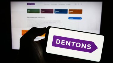 Stuttgart, Almanya - 08-24-2023: Küresel hukuk şirketi Dentons 'un logosunun bulunduğu cep telefonu sahibi kişi iş sayfasının önünde ekranda. Telefon ekranına odaklan.