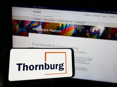 Stuttgart, Germany - 08-25-2023: Person holding mobile phone with logo of company Thornburg Investment Management Inc. on screen in front of web page. Focus on phone display. clipart