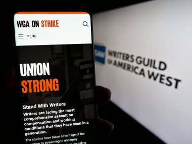 Stuttgart, Germany - 08-26-2023: Person holding smartphone with web page of labor union Writers Guild of America West (WGAW) on screen with logo. Focus on center of phone display. clipart