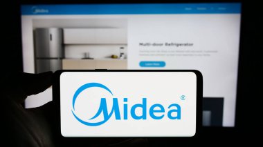 Stuttgart, Germany - 08-28-2023: Person holding cellphone with logo of Chinese home appliances company Midea Group on screen in front of business webpage. Focus on phone display. clipart