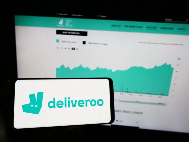 Stuttgart, Germany - 08-28-2023: Person holding mobile phone with logo of food delivery company Deliveroo Holdings plc on screen in front of web page. Focus on phone display. clipart