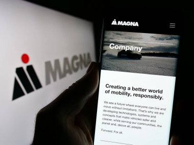 Stuttgart, Almanya - 08-29-2023: Otomotiv şirketi Magna International Inc. 'in web sitesi ile birlikte logo önünde cep telefonu tutan kişi. Telefon ekranının merkezine odaklan.