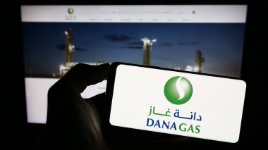 Stuttgart, Germany - 08-29-2023: Person holding mobile phone with logo of Emirati petroleum company Dana Gas PJSC on screen in front of business web page. Focus on phone display. clipart