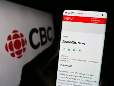 Stuttgart, Germany - 08-30-2023: Person holding smartphone with webpage of Canadian Broadcasting Corporation (CBC) on screen in front of logo. Focus on center of phone display. clipart