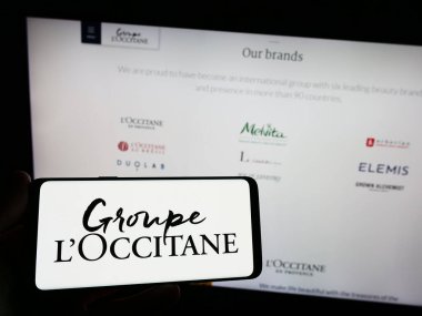 Stuttgart, Germany - 08-30-2023: Person holding smartphone with logo of French cosmetics company Groupe L'Occitane on screen in front of website. Focus on phone display. clipart