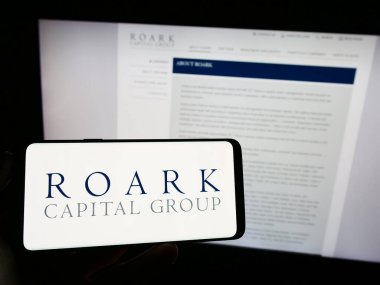 Stuttgart, Germany - 09-02-2023: Person holding mobile phone with logo of American company Roark Capital Management LLC on screen in front of web page. Focus on phone display. clipart