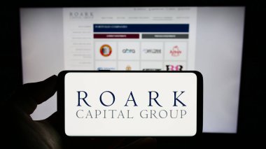 Stuttgart, Almanya - 09-02-2023: ABD 'li Roark Capital Management LLC logosuna sahip cep telefonu sahibi kişi iş sayfasının önünde ekranda. Telefon ekranına odaklan.