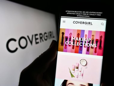 Stuttgart, Germany - 09-05-2023: Person holding cellphone with webpage of US cosmetics company CoverGirl on screen in front of business logo. Focus on center of phone display. clipart