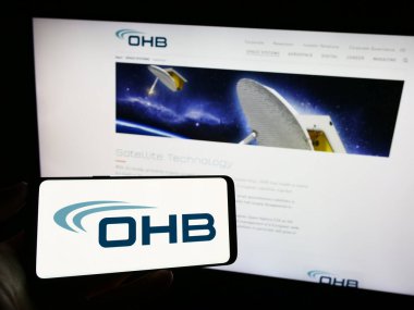 Stuttgart, Almanya - 09-10-2023: Alman havacılık şirketi OHB SE 'nin logosunu taşıyan cep telefonu sahibi kişi iş sayfasının önünde ekranda. Telefon ekranına odaklan.