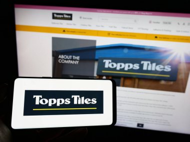 Stuttgart, Germany - 07-27-2023: Person holding smartphone with logo of British retail company Topps Tiles plc on screen in front of website. Focus on phone display. clipart