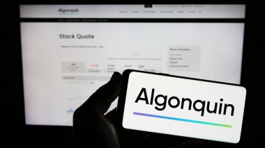 Stuttgart, Germany - 07-28-2023: Person holding mobile phone with logo of Canadian company Algonquin Power Utilities Corp. on screen in front of web page. Focus on phone display. clipart