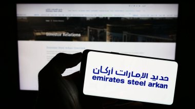 Stuttgart, Germany - 07-30-2023: Person holding smartphone with logo of company Emirates Steel Arkan (UAE) on screen in front of website. Focus on phone display. clipart