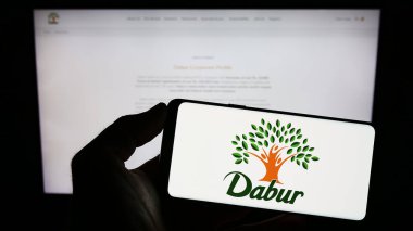 Stuttgart, Germany - 07-25-2023: Person holding smartphone with logo of Ayurveda company Dabur India Limited on screen in front of website. Focus on phone display. clipart