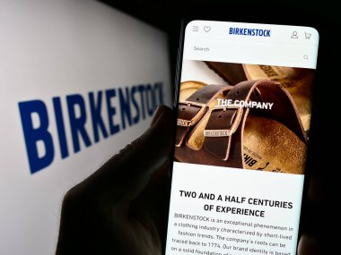 Stuttgart, Almanya - 07-24-2023: Ayakkabı şirketi Birkenstock Group B.V. Co. KG 'nin web sitesi ile cep telefonu tutan kişi logolu ekranda. Telefon ekranının merkezine odaklan.