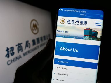 Stuttgart, Almanya - 07-21-2023: Nakliye şirketi China Merchants Group Limited 'un internet sayfası ile cep telefonu tutan kişi logolu ekranda. Telefon ekranının merkezine odaklan.