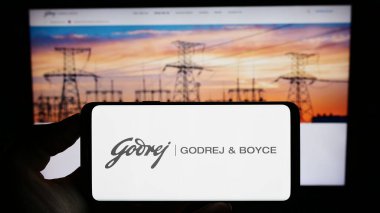 Stuttgart, Germany - 07-21-2023: Person holding cellphone with logo of Indian company Godrej and Boyce Mfg. Co. Ltd. on screen in front of webpage. Focus on phone display. clipart