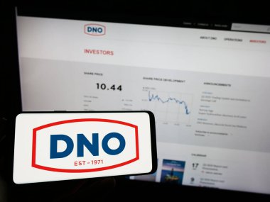 Stuttgart, Germany - 07-21-2023: Person holding mobile phone with logo of Norwegian oil and gas company DNO ASA on screen in front of business web page. Focus on phone display. clipart