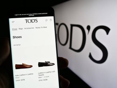 Stuttgart, Germany - 07-15-2023: Person holding cellphone with webpage of Italian shoe company Tod's SpA on screen in front of business logo. Focus on center of phone display. clipart