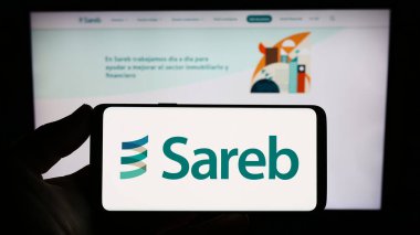 Stuttgart, Germany - 07-15-2023: Person holding smartphone with logo of Spanish bad bank Sareb on screen in front of website. Focus on phone display. clipart