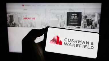 Stuttgart, Germany - 07-15-2023: Person holding mobile phone with logo of real estate company Cushman Wakefield plc on screen in front of web page. Focus on phone display. clipart