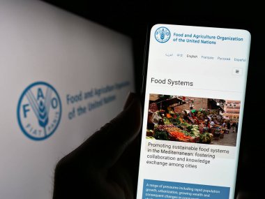 Stuttgart, Germany - 07-11-2023: Person holding cellphone with website of UN agency Food and Agriculture Organization (FAO) on screen with logo. Focus on center of phone display. clipart