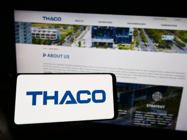 Stuttgart, Germany - 07-08-2023: Person holding mobile phone with logo of company Truong Hai Auto Corporation (THACO) on screen in front of web page. Focus on phone display. clipart