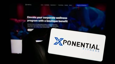 Stuttgart, Germany - 07-08-2023: Person holding smartphone with logo of US franchise company Xponential Fitness Inc. on screen in front of website. Focus on phone display. clipart