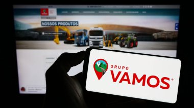 Stuttgart, Germany - 07-06-2023: Person holding cellphone with logo of Brazilian truck rental company Grupo Vamos on screen in front of business webpage. Focus on phone display. clipart