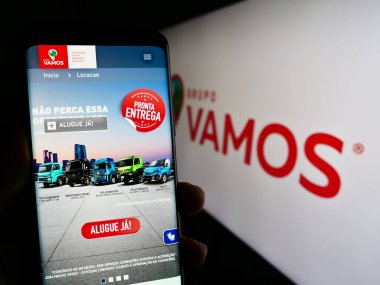 Stuttgart, Germany - 07-06-2023: Person holding cellphone with webpage of Brazilian truck rental company Grupo Vamos on screen in front of logo. Focus on center of phone display. clipart