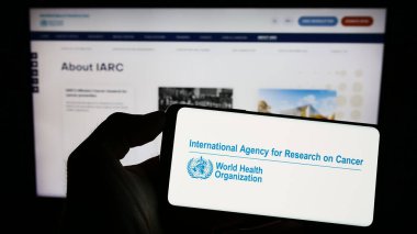 Stuttgart, Almanya - 07-04-2023: İnternet sayfası önünde Uluslararası Kanser Araştırmaları Dairesi (IARC) logosuyla cep telefonu tutan kişi. Telefon ekranına odaklan.