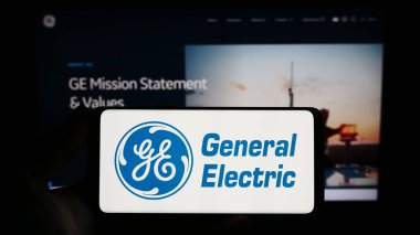Stuttgart, Almanya - 07-01-2023: İnternet sayfası önünde üzerinde Amerikan holdingi General Electric Company (GE) logosu bulunan cep telefonu sahibi kişi. Telefon ekranına odaklan.